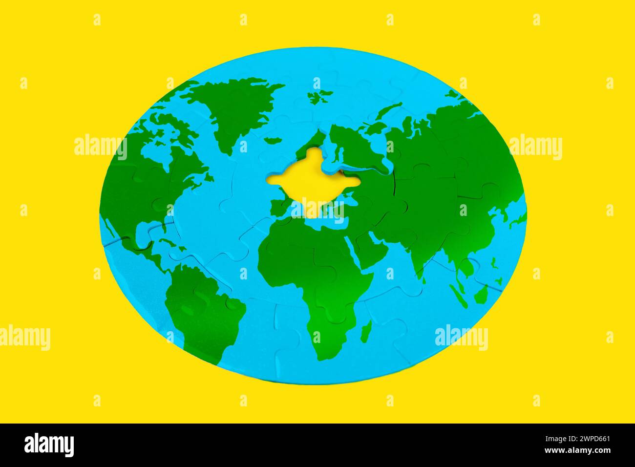 Round jigsaw puzzle depicts a world map with green continents and blue oceans on a yellow background. The puzzle is incomplete, with one piece missing Stock Photo
