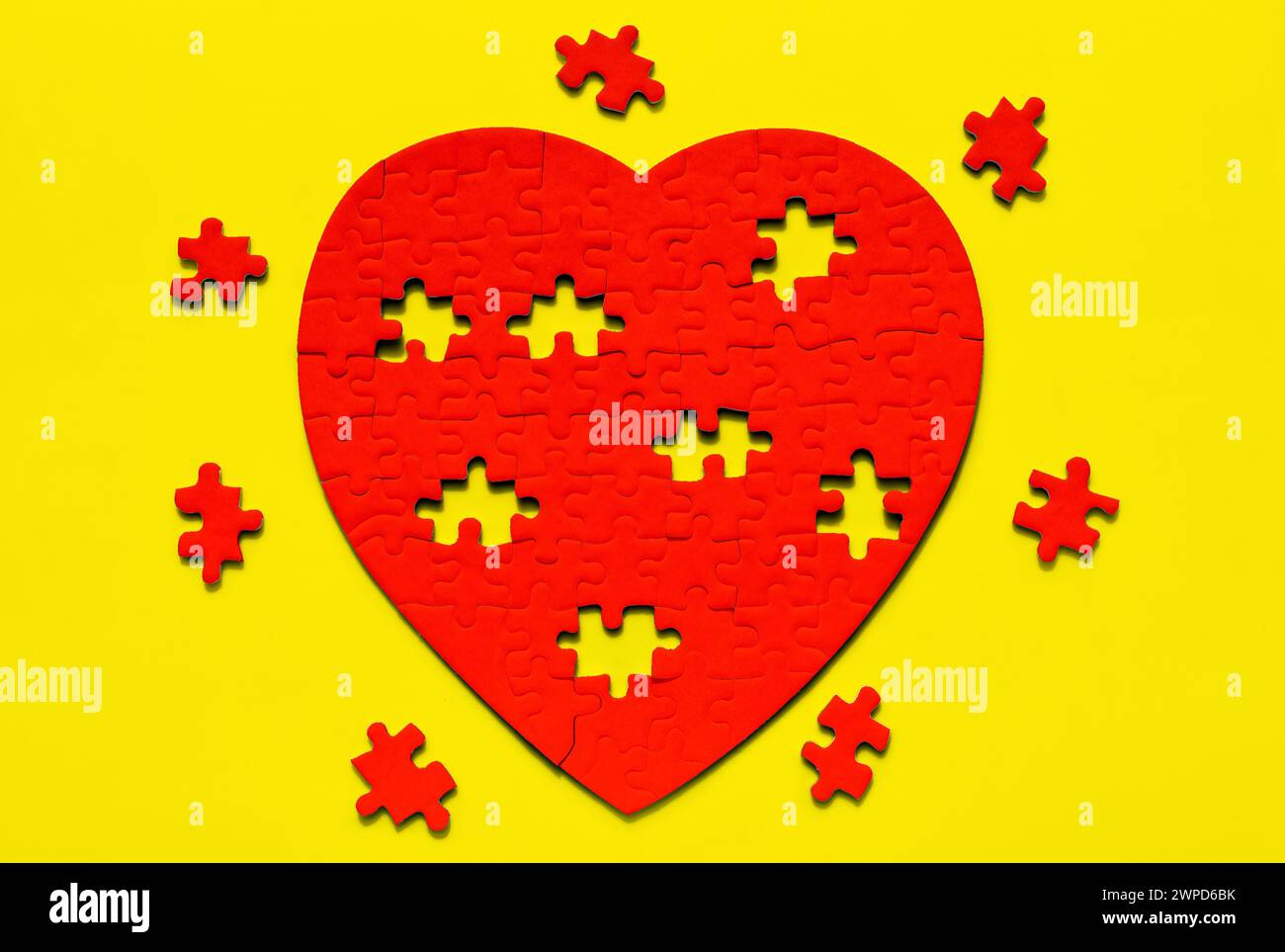 Red heart-shaped puzzle resting on a yellow backdrop, with missing pieces arranged around it. Creative Valentine's Day background. Stock Photo