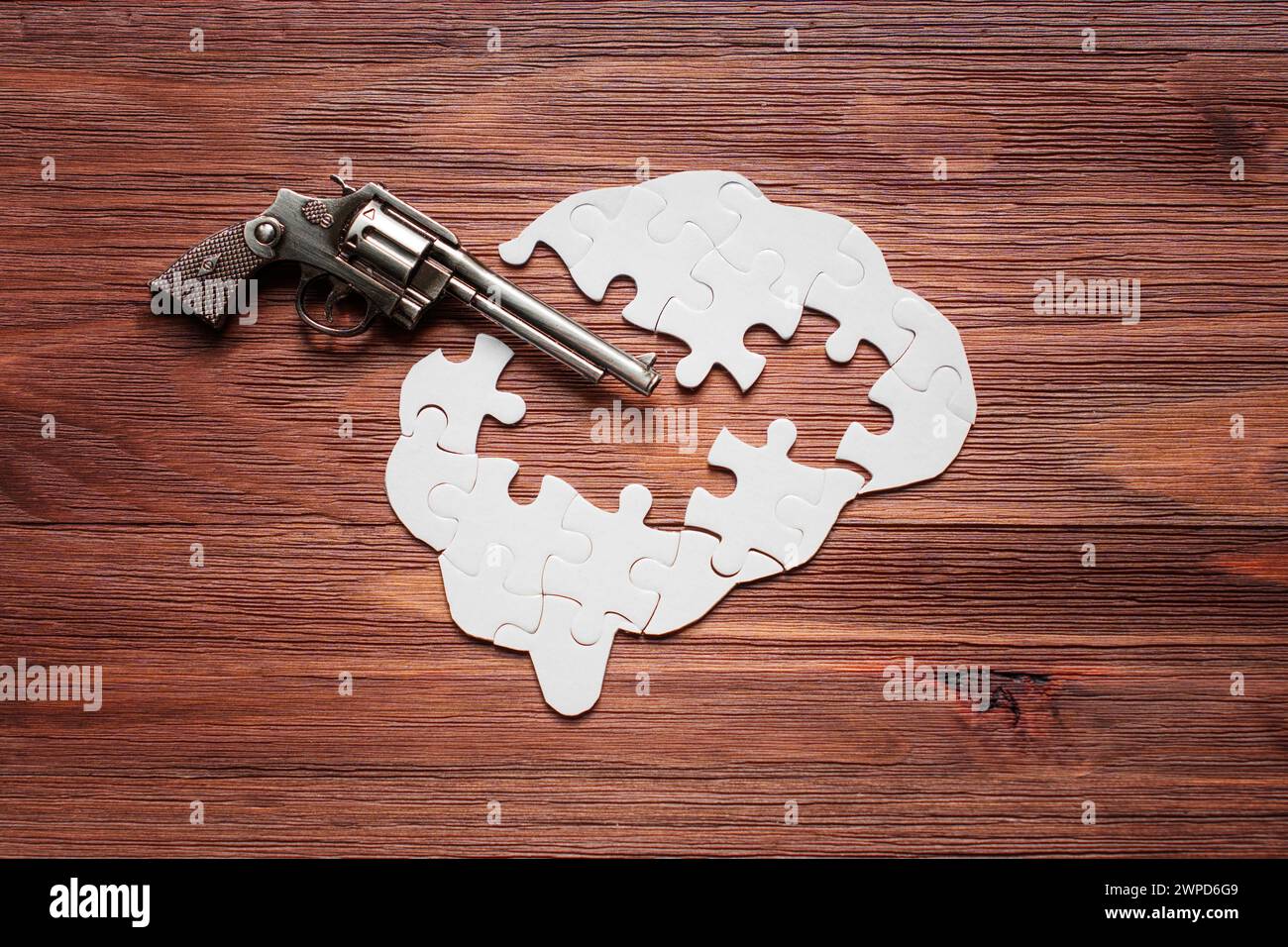 Revolver inserted into a human brain puzzle, set against a textured wooden backdrop. With several missing puzzle pieces, the composition metaphoricall Stock Photo