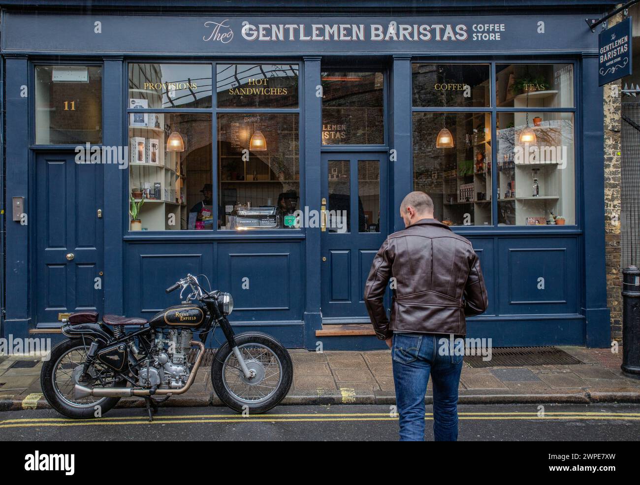 Rear view of man in leather jacket  walking towards coffee shop with classic motorcycle parked in front of cafe . Stock Photo