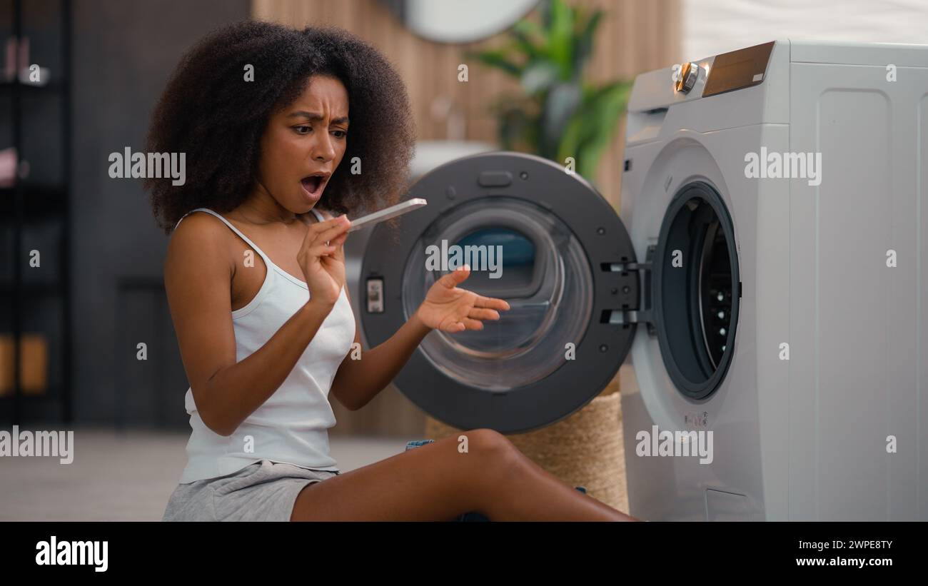 African American girl ethnic lady biracial woman housewife home laundry clothes open washing machine housekeeping take clean wet jeans pants find Stock Photo