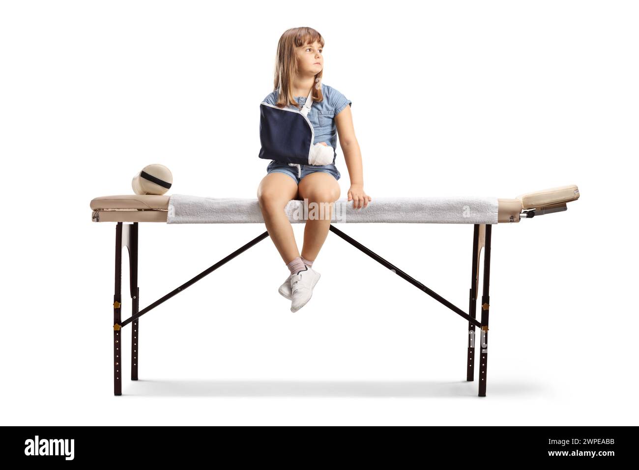 Little girl with a broken arm wearing a sling and sitting on a physical therapy bed isolated on