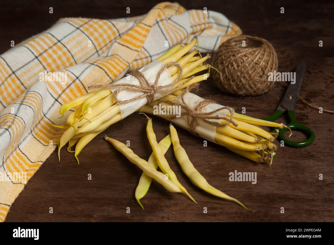 Two bunch and several pods of raw yellow pods of haricot with yellow kitchen towel, scissors and ball of thread on wooden background. French beans or Stock Photo