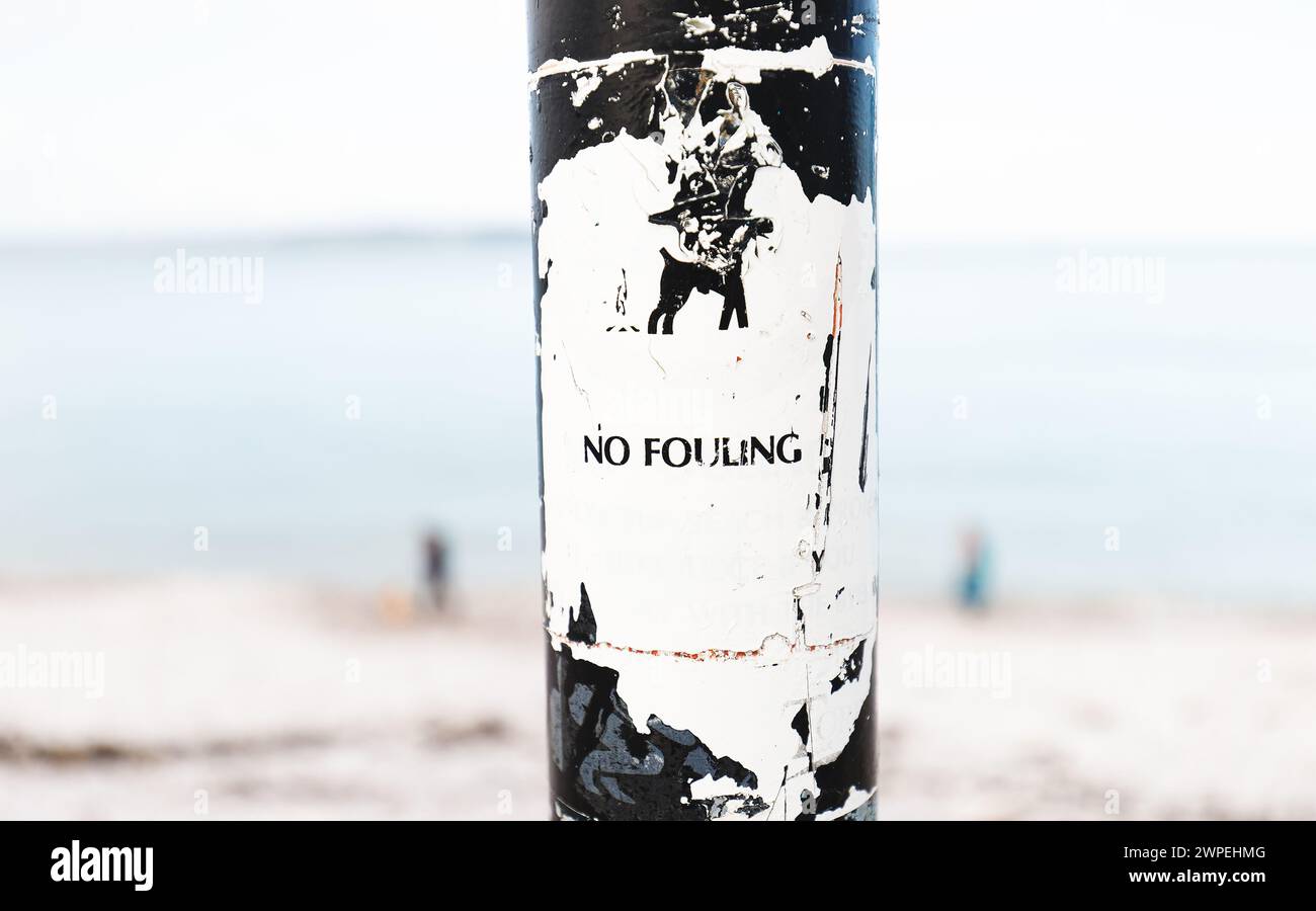 Dog Fouling Notice on Post Worn and defaced with image of dog with blurred background of beach with two walkers Stock Photo