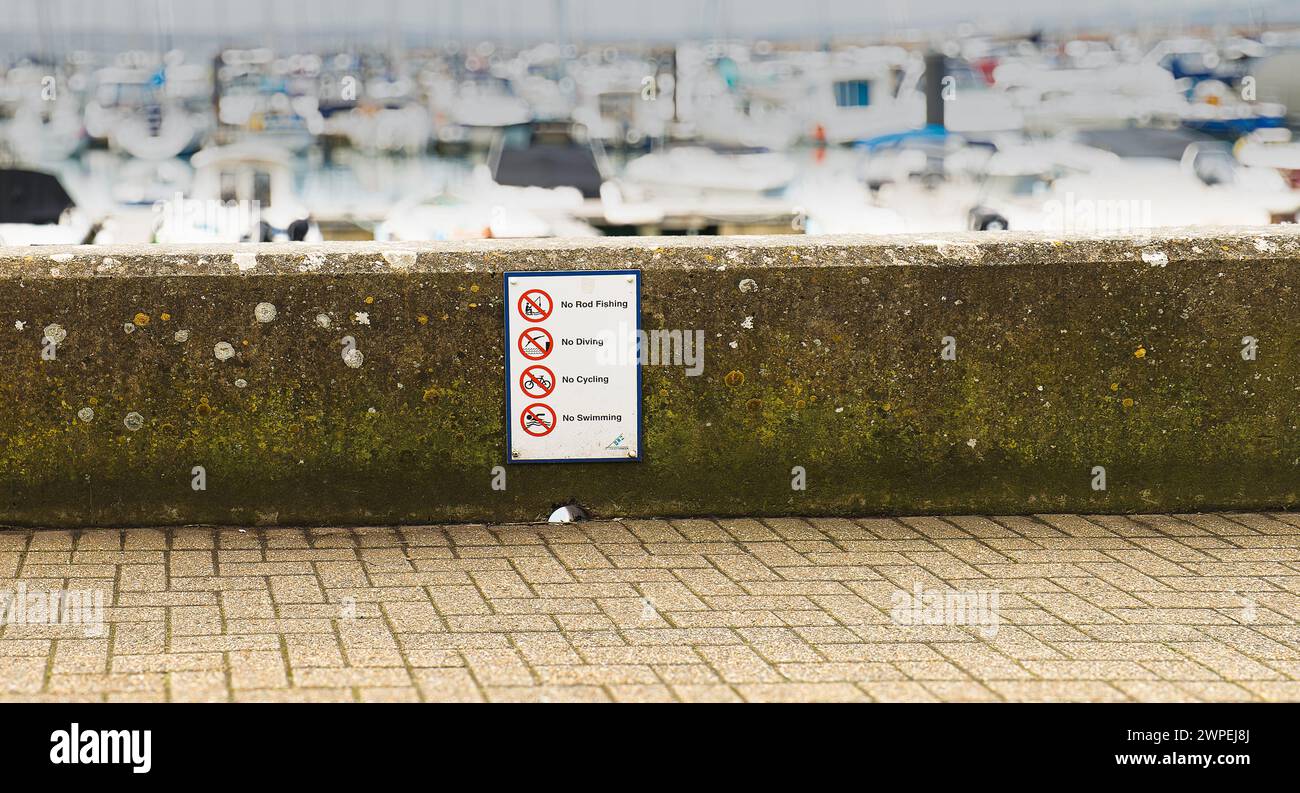Prohibition sign on harbour wall saying No Fishing, Diving, Cycling or Swimming With blurred boats behind the wall and block paving pavement sidewalk Stock Photo