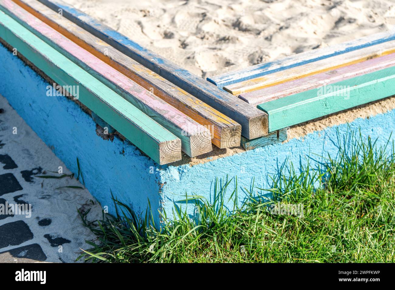 Concrete barrier with colorful wooden bars serving as seats, surrounding a children's sandpit near lush lawn on sunny day Stock Photo