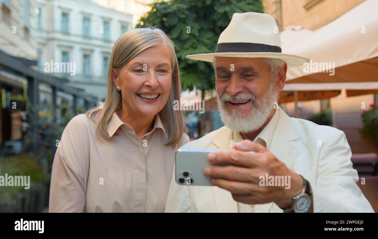 Happy smiling Caucasian senior elderly couple walking taking selfie laughing talking gadget mobile phone together city street outdoors. Old man woman Stock Photo