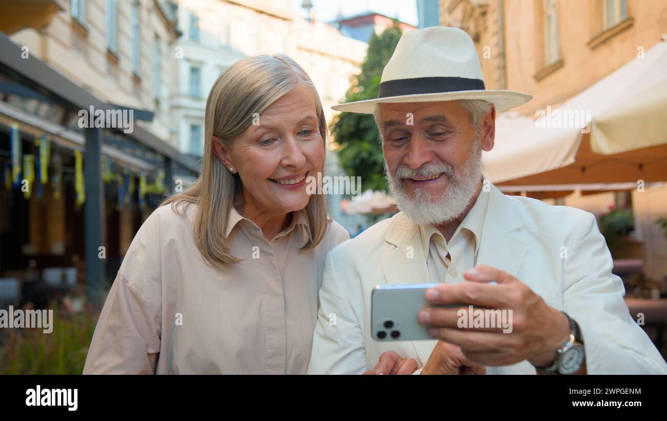 Happy smiling Caucasian senior elderly couple walking taking selfie laughing talking gadget mobile phone together city street outdoors. Old man woman Stock Photo