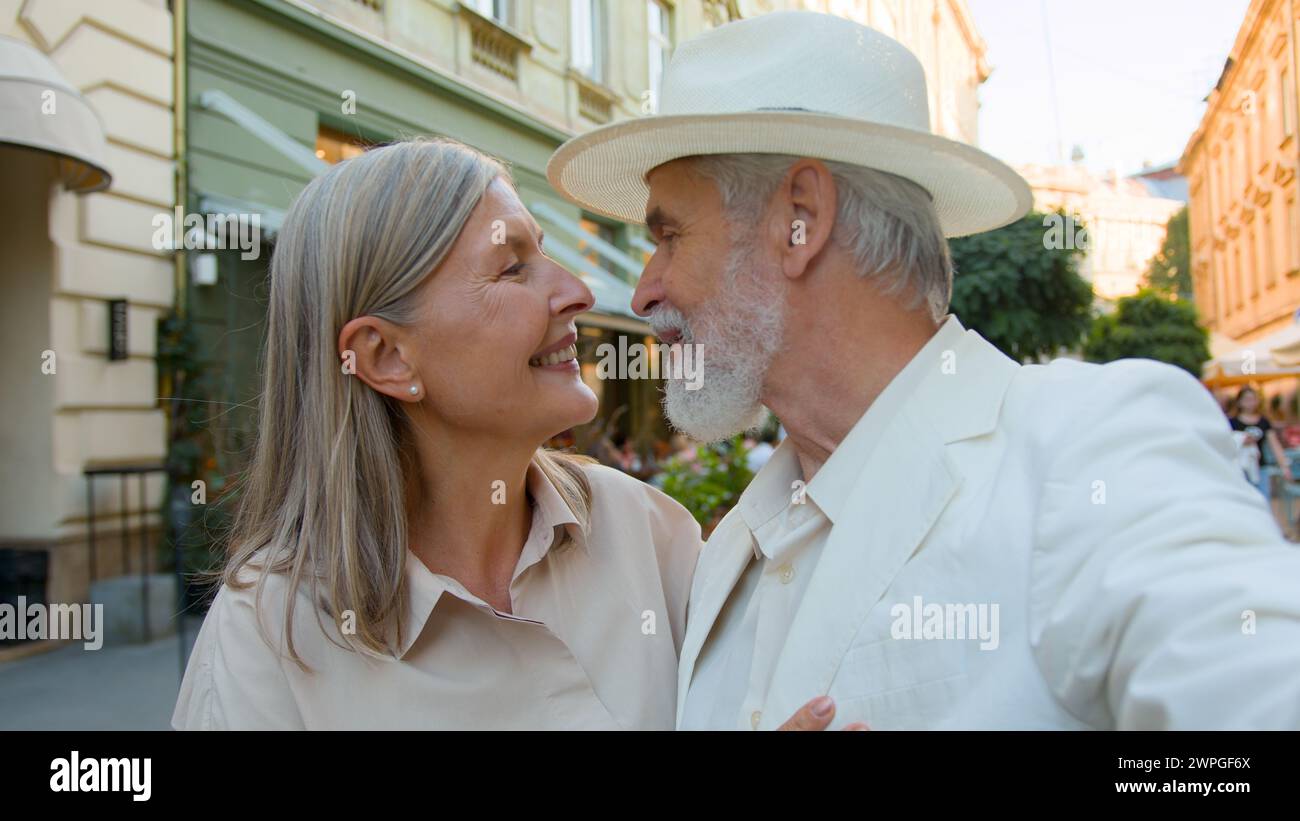 Happy elderly Caucasian tourists couple senior man woman taking selfie streaming video romantic kissing mobile phone modern technologies outside city Stock Photo