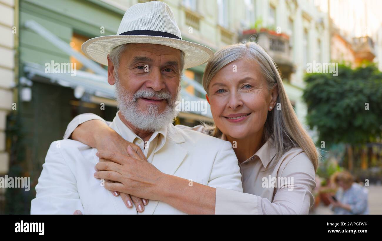 Portrait joyful aged Caucasian happy family elderly wide smiling pensioners woman hugging embrace man looking at camera happiness outdoors city Stock Photo