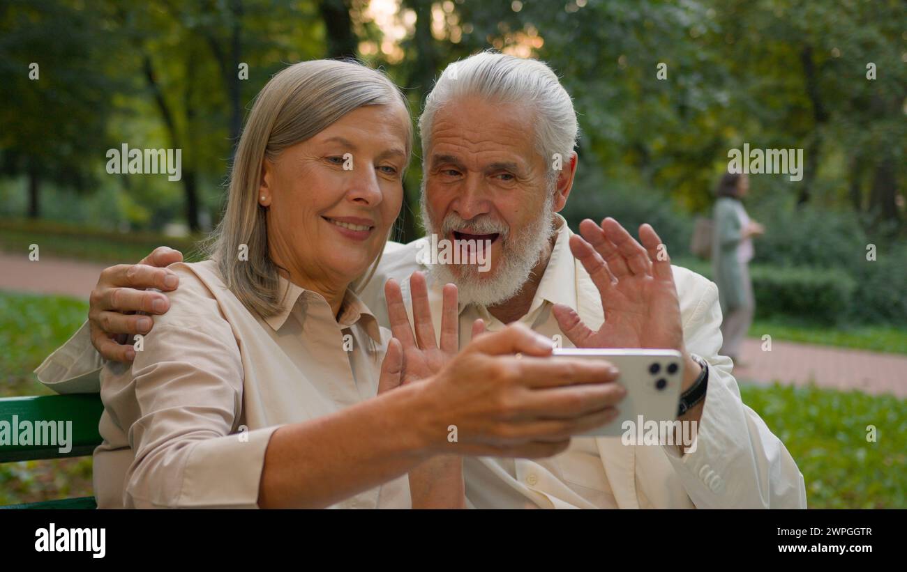 Close up portrait smiling Caucasian middle aged retired family couple talking relaxing using smartphone outdoors city park. Happy grandparents online Stock Photo