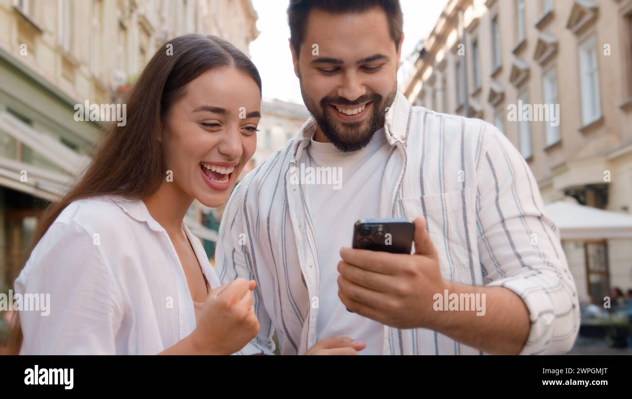 Caucasian couple happy woman man rejoices winning victory lucky expression together joy using mobile phone cellphone high-five exclamation success Stock Photo