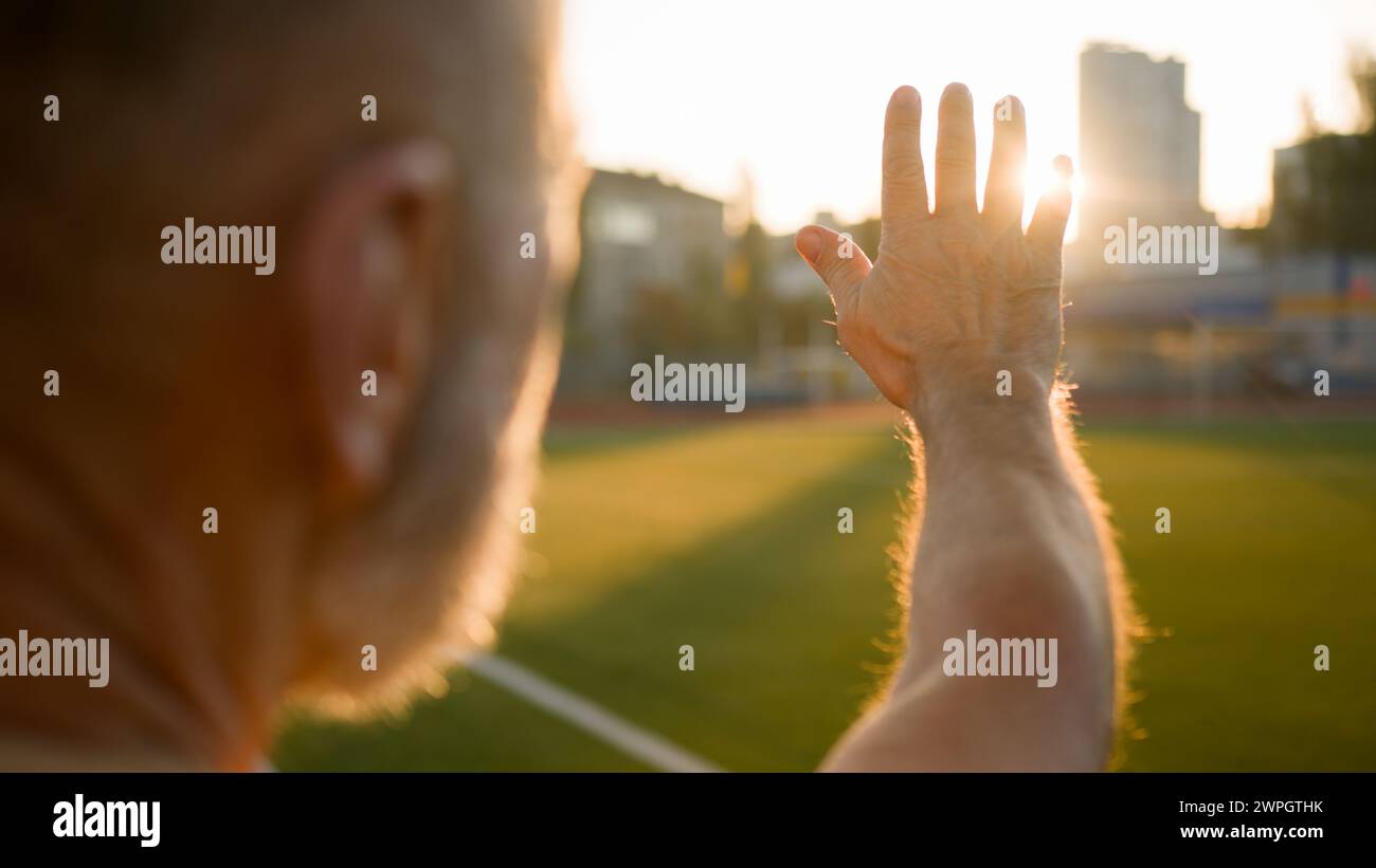 Close up old man grandfather looking at sun rays put hand out sunset warmth fresh air training stadium city outside good weather calm peaceful sports Stock Photo