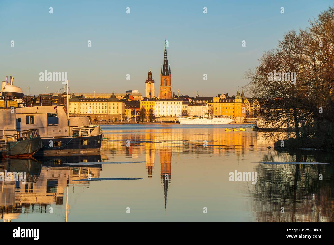 Riddarholmen church reflection in the lake at sunset. Stockholm, Sweden. Blue sky, boats in foreground, late winter. Yellow buildings. Stock Photo