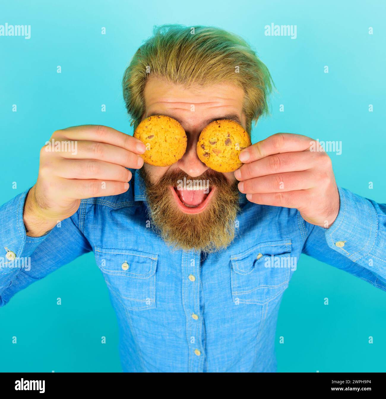 Bearded man covered eyes with freshly baked oatmeal cookies. Tasty sweet snack. Handsome male eating chocolate cookies biscuit. Breakfast or lunch Stock Photo