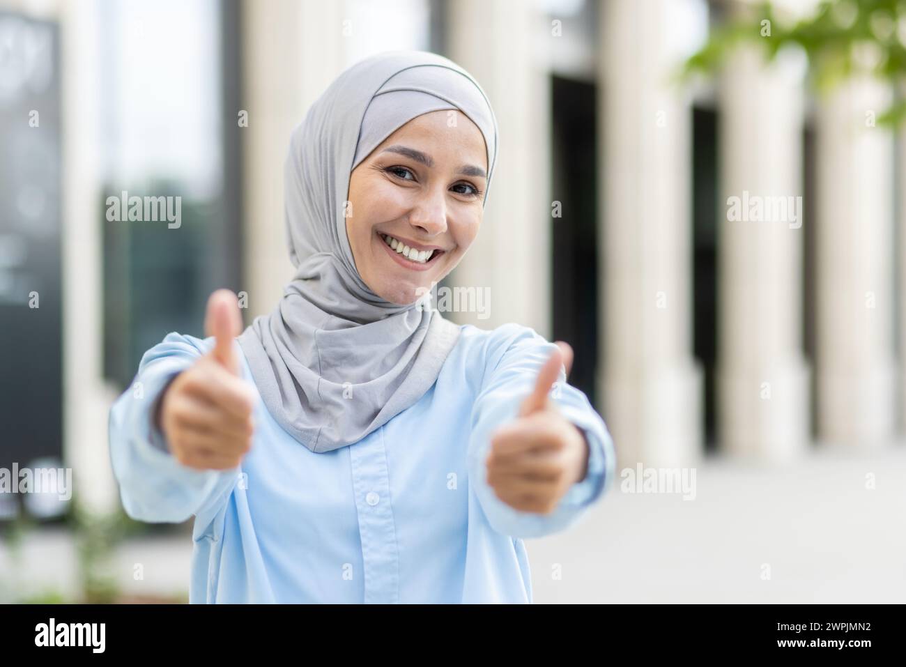 A cheerful, young woman wearing a hijab gives a double thumbs up with a broad, confident smile, signaling approval and success. Stock Photo