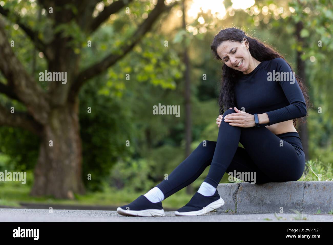 Active woman outdoors holding her knee with a pained expression, indicating a sports injury during workout. Stock Photo