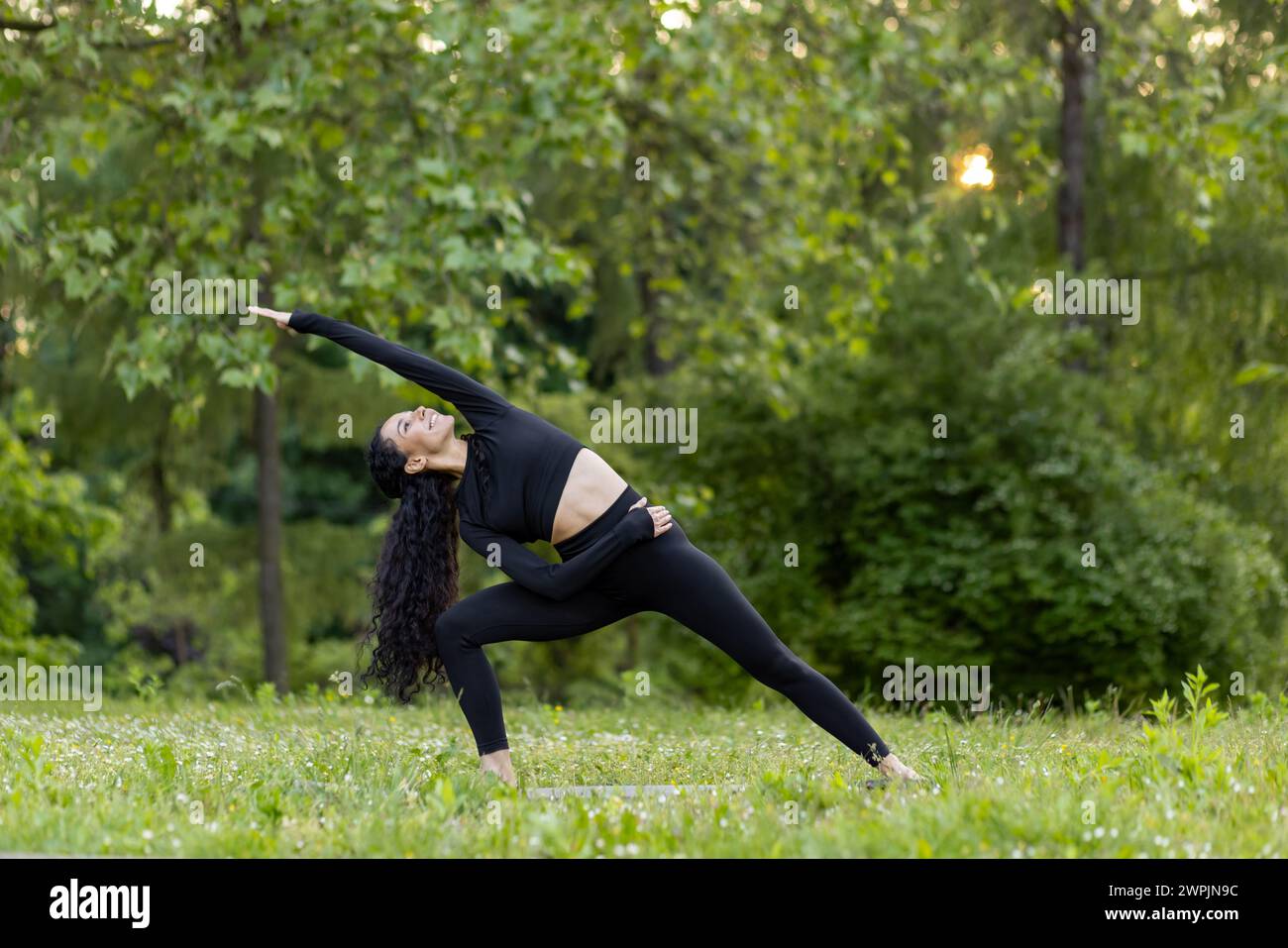 An active young woman performs a yoga pose outdoors, surrounded by vibrant green trees and the glow of the sunset. Stock Photo