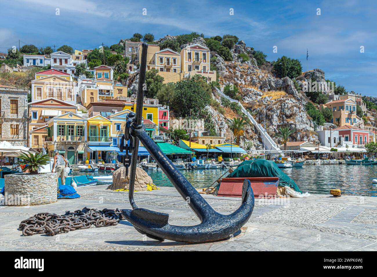 SYMI, GREECE - JULY 3, 2022: Promenade of the city with metal anchor sculpture and multi-colored typical houses. Stock Photo