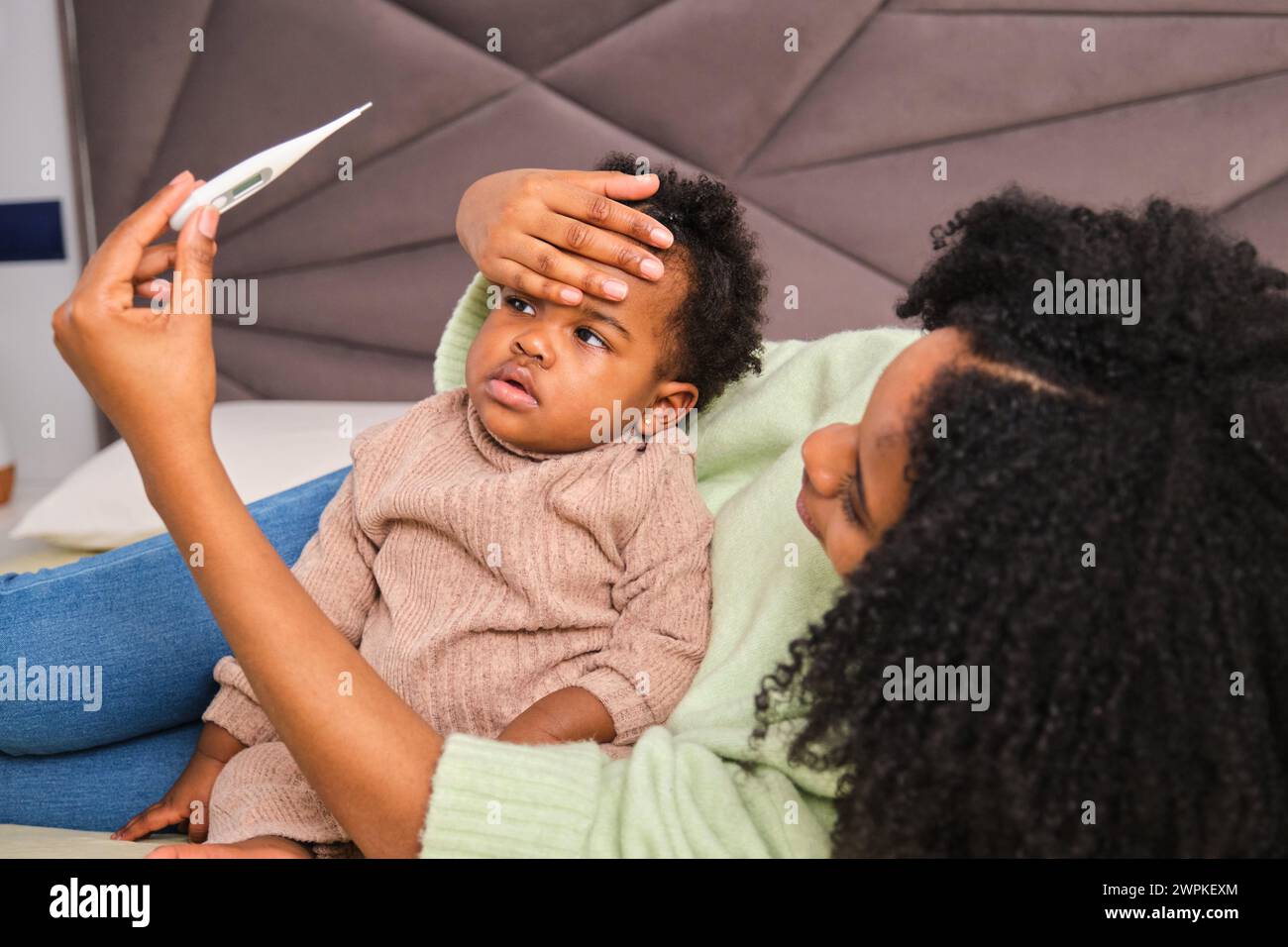 Cuban mother measuring her toddler daughter's fever on bed. Stock Photo