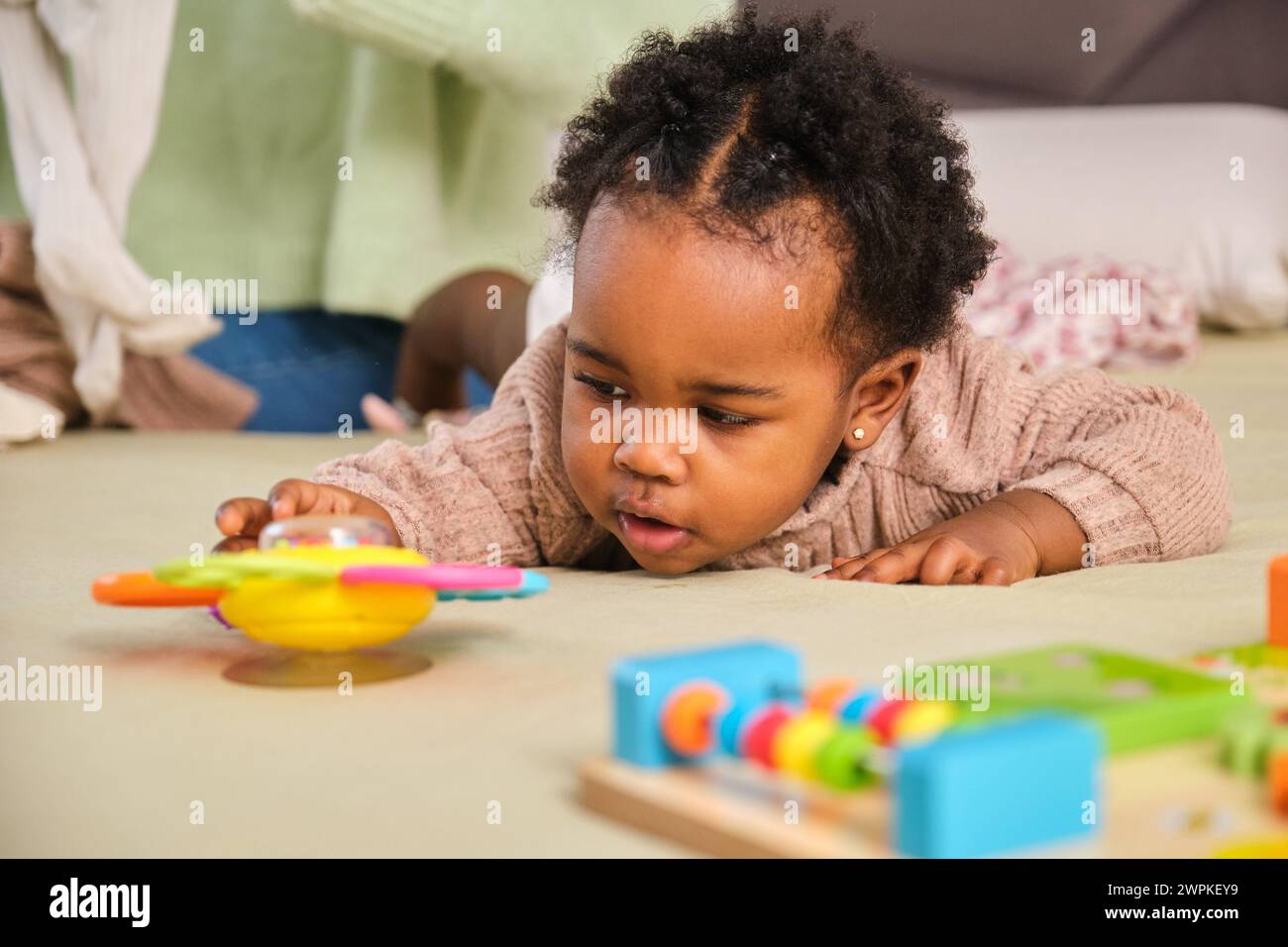 Cuban toddler baby girl trying to reach a toy playing on bed at home. Stock Photo