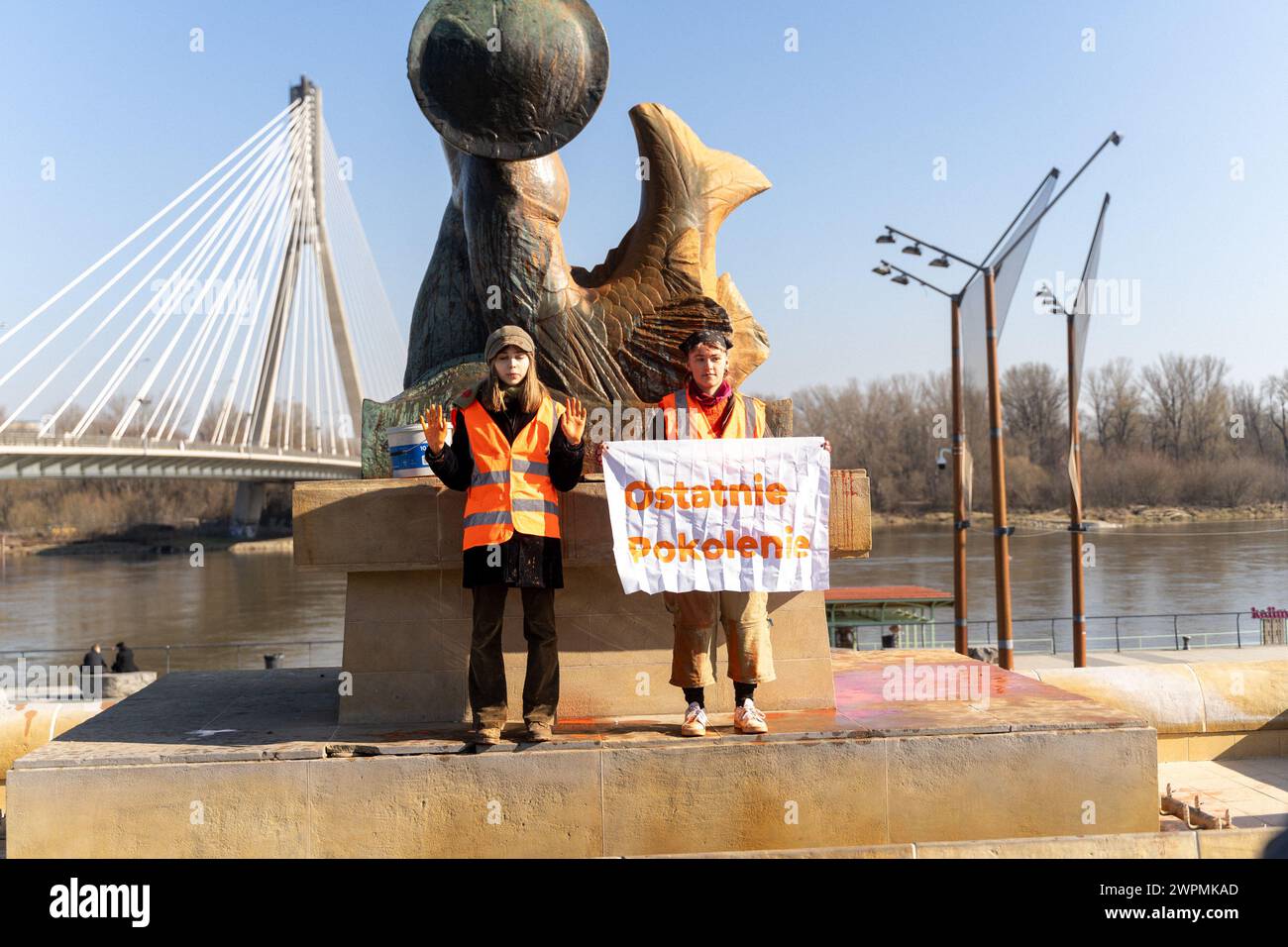 Climate activists stand on the monument of Warsaw mermaid doused with paint, one of the activists holds a banner saying 'The Last Generation' during the demonstration. Warsaw's 'Syrenka' was doused with orange paint as part of a protest by the Last Generation group, which demands effective action to combat the climate crisis. Two women carried out an action on the Vistula Boulevards, voicing the campaign's demands, and then were detained by the police. The action took place on Women's Day to emphasize the role of women in the fight for the environment. The group demands greater investment in p Stock Photo