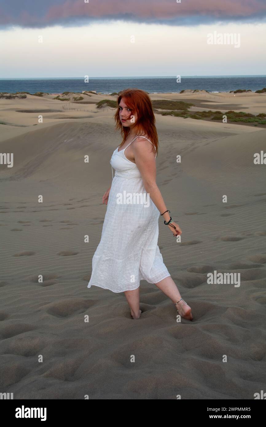A young girl stands in a white dress in the sand dunes with sky and clouds in the late afternoon Stock Photo