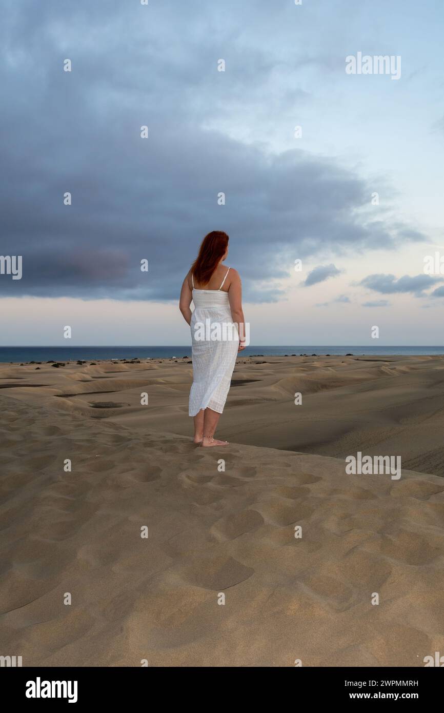 A young girl stands in a white dress in the sand dunes with sky and clouds in the late afternoon Stock Photo