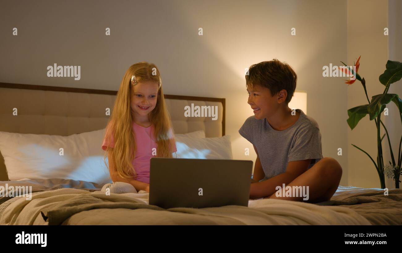 Laughing Caucasian happy children brother sister kids girl boy looking laptop screen in bedroom on bed evening watching cartoon video online using Stock Photo