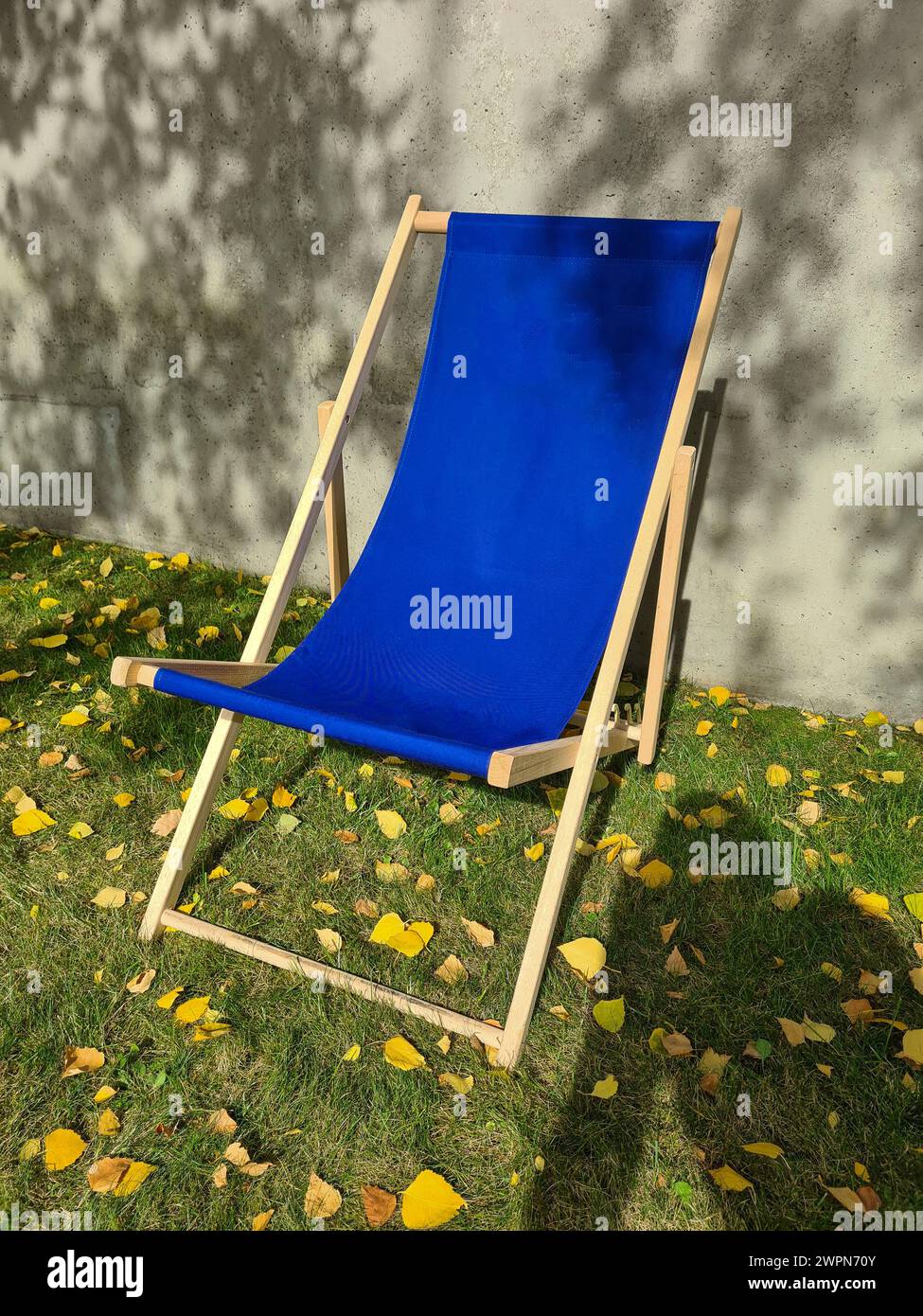 A blue deck chair stands in the sunlight on a green lawn in front of a light gray concrete wall with shadows of leaves Stock Photo