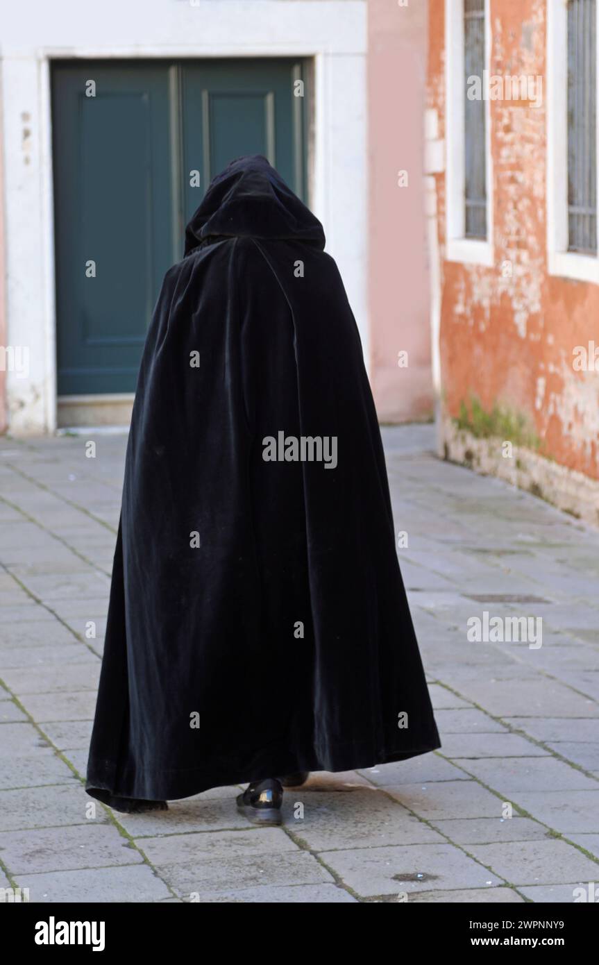 figure in a hood walks through a city alley wearing a worn black tabard as a cloak Stock Photo