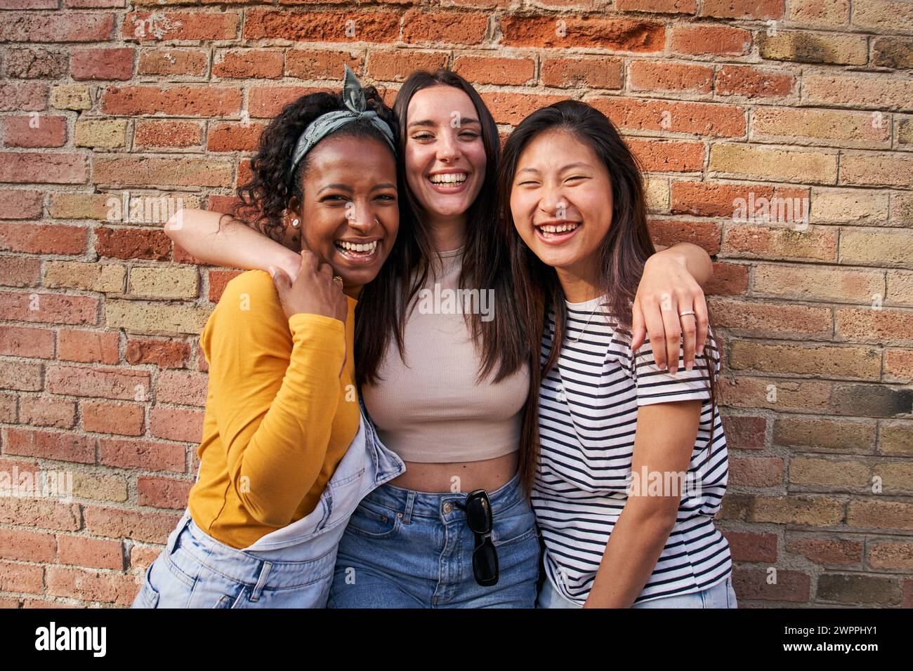 Portrait of three multiracial cheerful young women standing hugging each other brick wall outdoors. Stock Photo