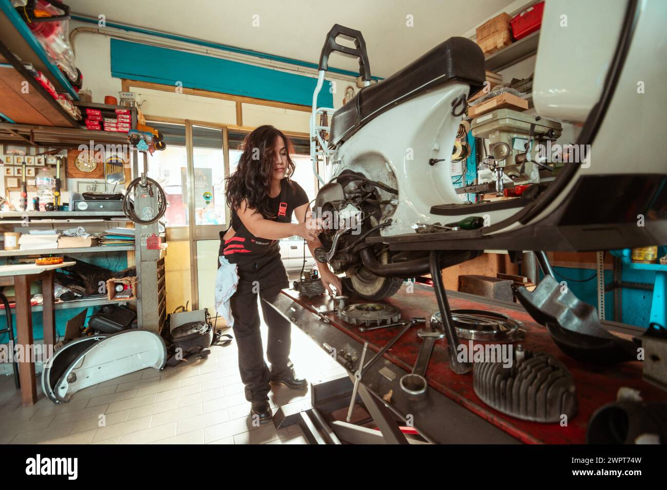 A woman mechanic works on a vintage scooter in a cluttered workshop, wearing black overall and shirt, latino female in traditional masculine jobs Stock Photo