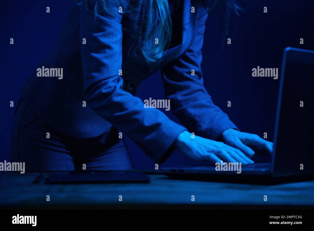 Business woman standing working on a laptop in the dark and illuminated by the screen. No face. Focus on hands. Blue color correction. Stock Photo