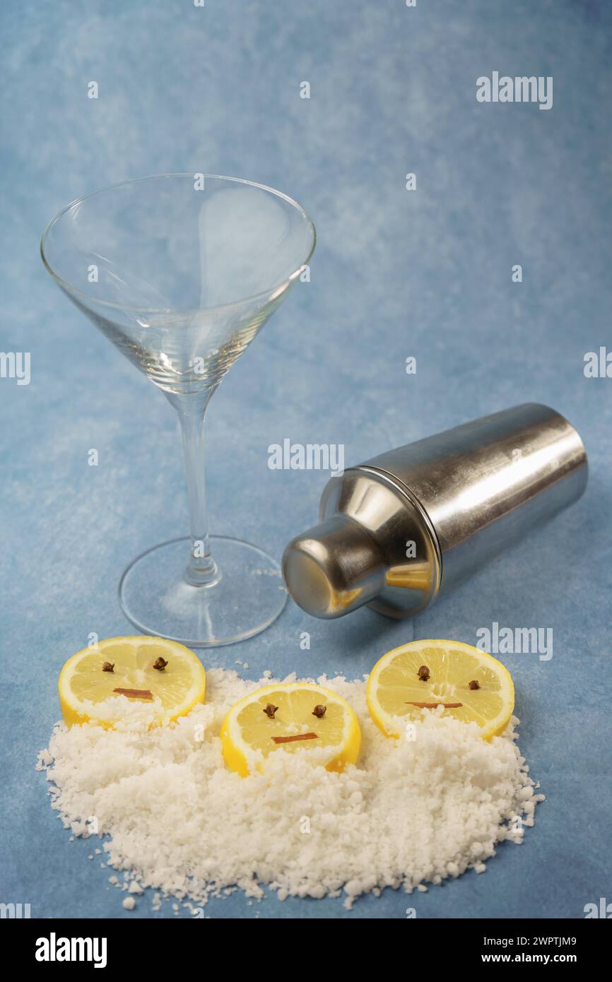 Lemon slices with cloves and cinnamon forming a smiley face on a pile of salt next to a cocktail shaker and a crystal glass with blue background with Stock Photo