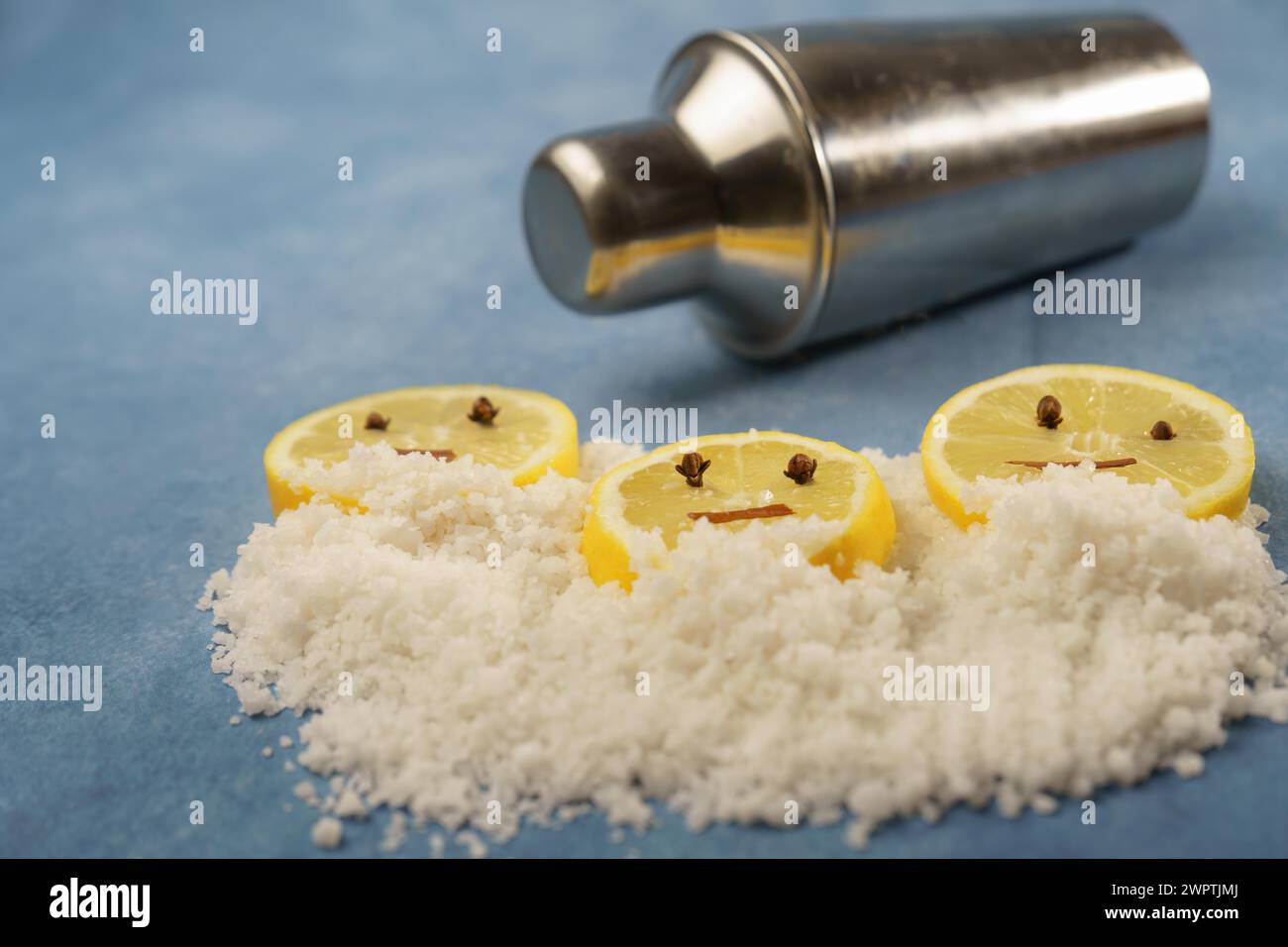 Lemon slices with cloves and cinnamon forming a smiley face on a pile of salt next to a shaker with blue background with copy space Stock Photo
