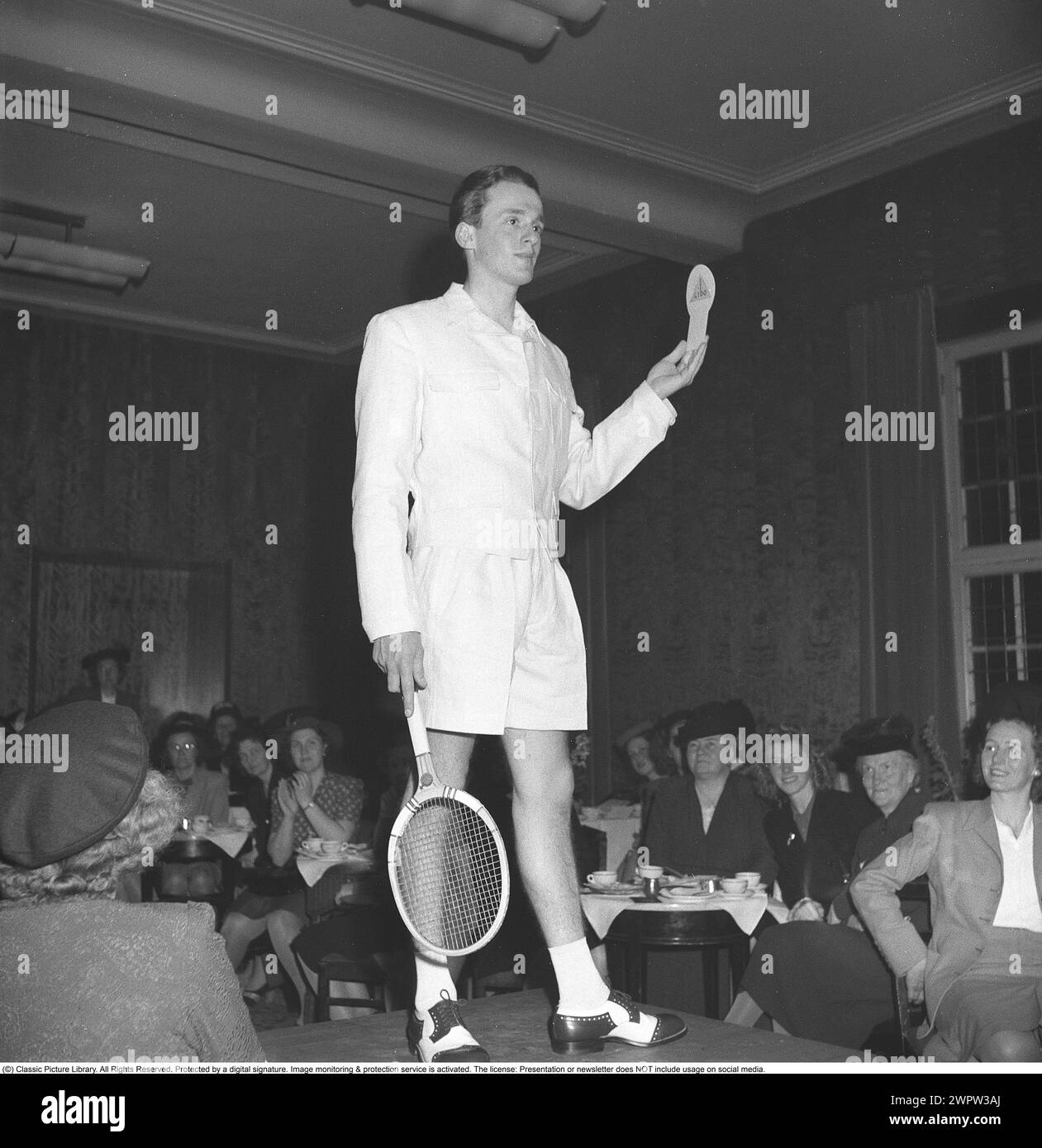 Fashion show in the 1940s. A young dark haired male model at a fashion show shows off a pair of trendy tennis pants and matching white jacket with a tennis racket in hand. The all-female audience views the garment and the man from top to bottom. 1946 Kristoffersson ref U1-1 Stock Photo