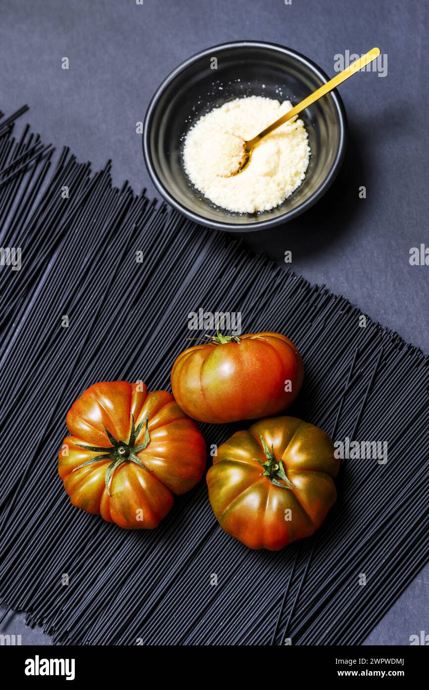 A trio of succulent ripe Raf tomatoes on black-dyed spaghetti, next to a black bowl filled with grated cheese with a golden spoon Stock Photo