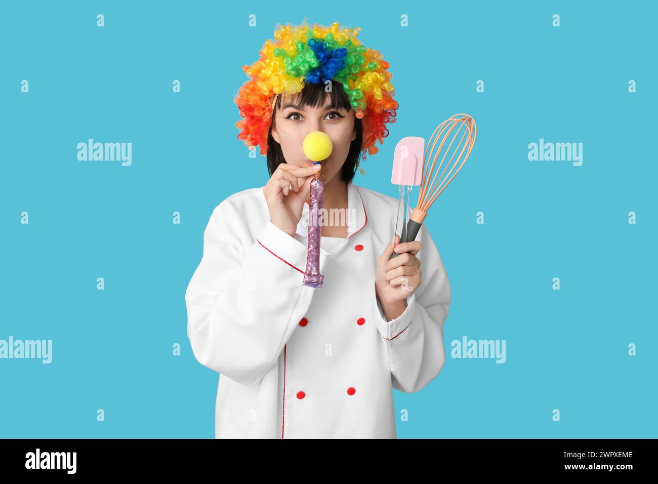 Young laughing female chef with party blower and kitchen utensils on blue background. Fool's day Stock Photo