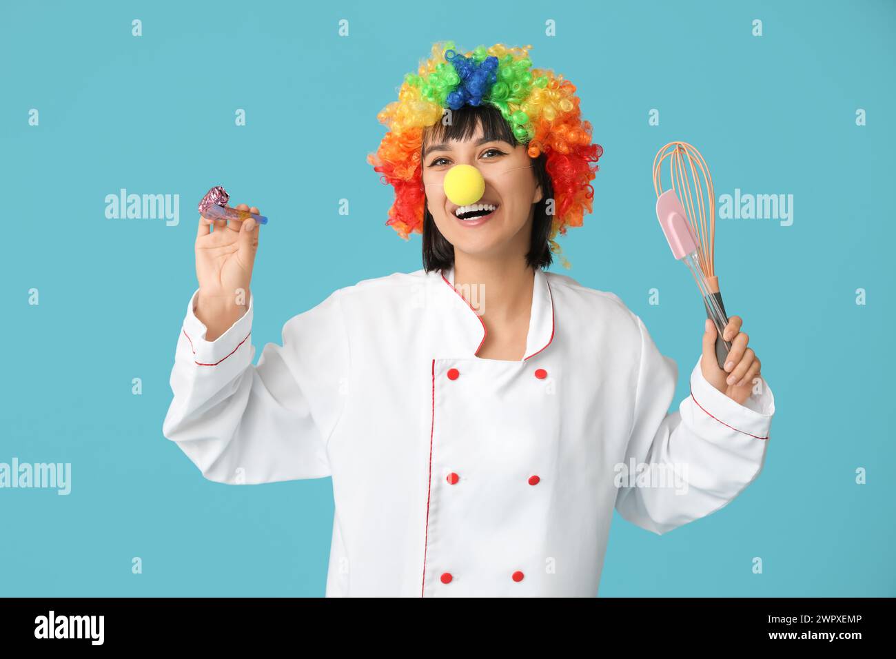Young laughing female chef with party blower and kitchen utensils on blue background. Fool's day Stock Photo