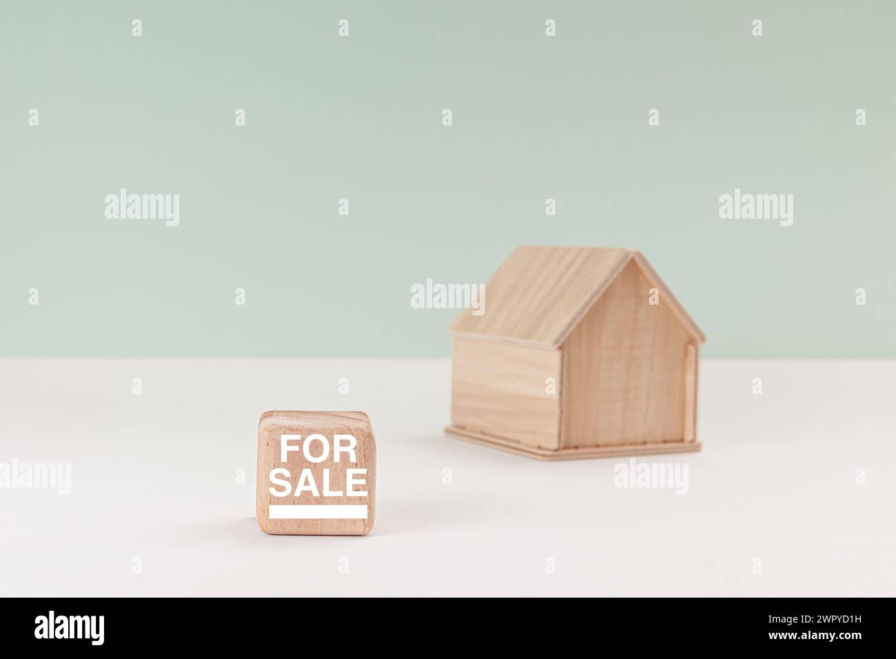 Simplistic wooden house model isolated on pale green background, with text For Sale on signboard. Stock Photo