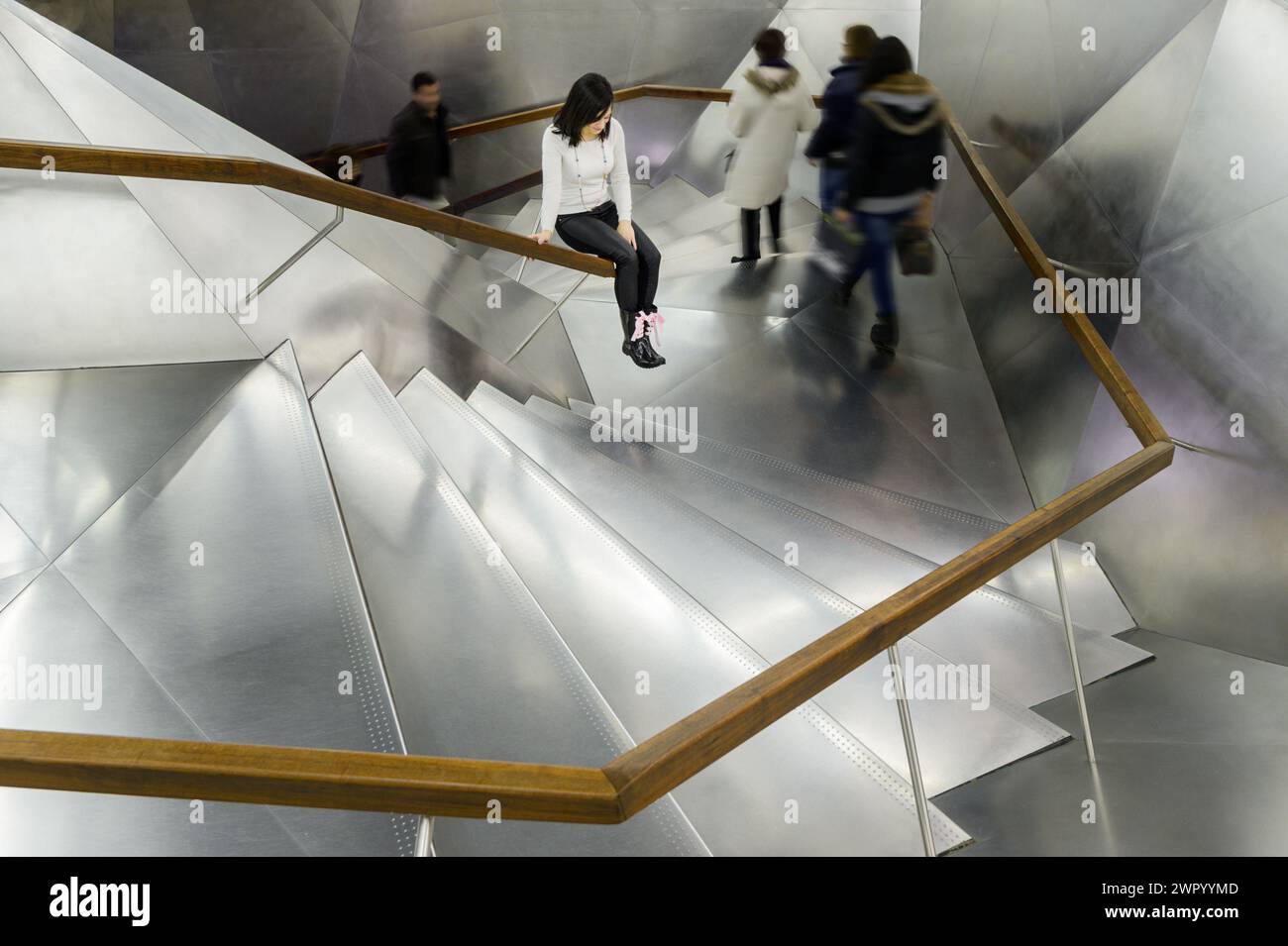 A girl sitting on a railing of modern aluminium stairs as people walk ...
