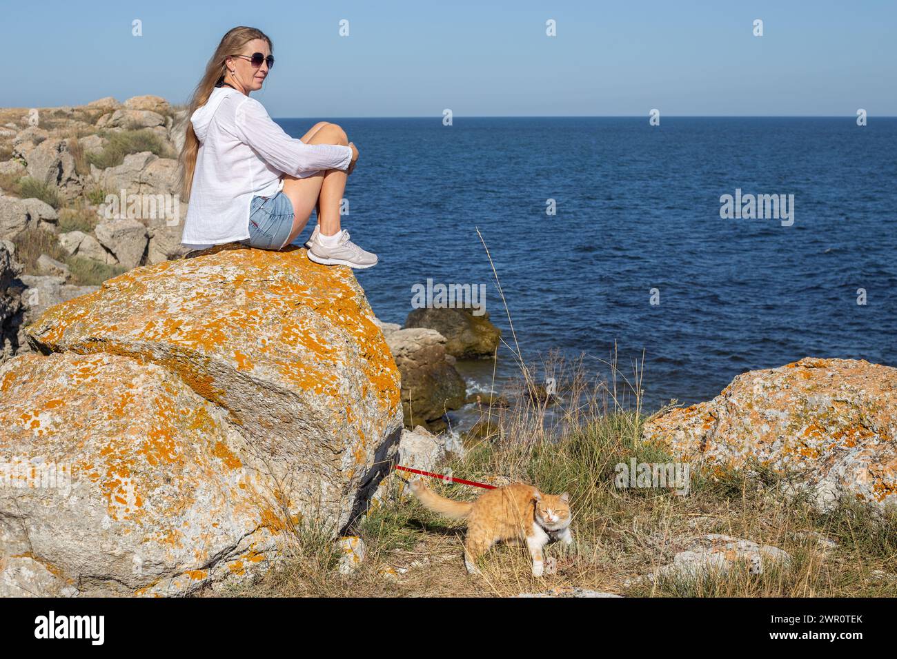 mature woman with long blond hair sits on a stone on the seashore, a cat on a leash walks nearby. Traveling and tourism with pets. Stock Photo