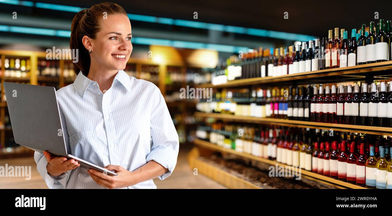 Woman sales manager of supermarket. Woman with laptop stands in front of wine bottles on shelves and smiling. Stock Photo