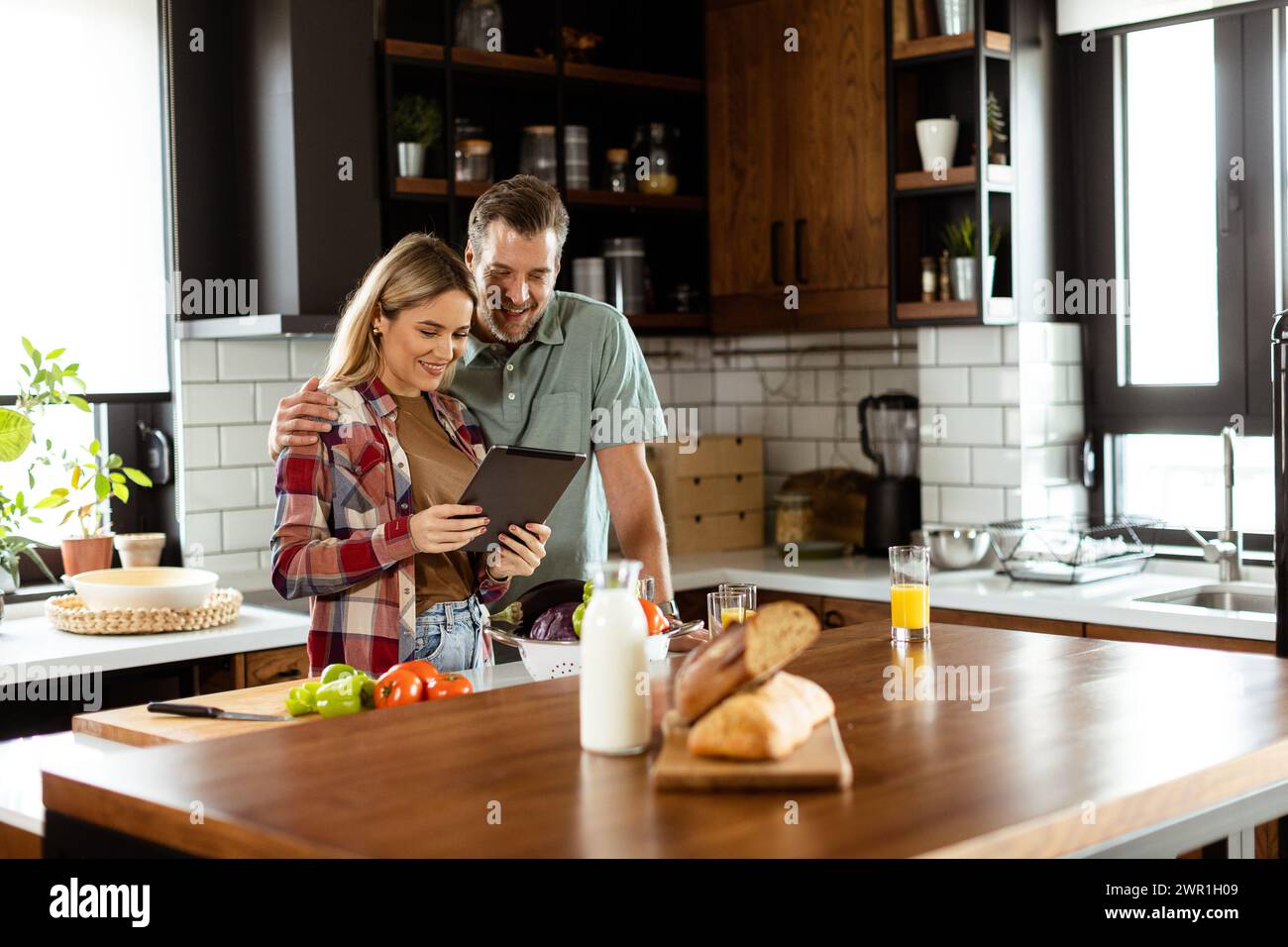 A cheerful couple stands in a well-lit kitchen, engrossed in a digital tablet among fresh ingredients Stock Photo