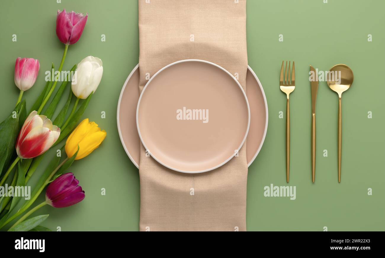 Festive place setting with beige napkin and spring flowers. Empty plates and gold cutlery on green background. Top view. Dining table in luxury restau Stock Photo