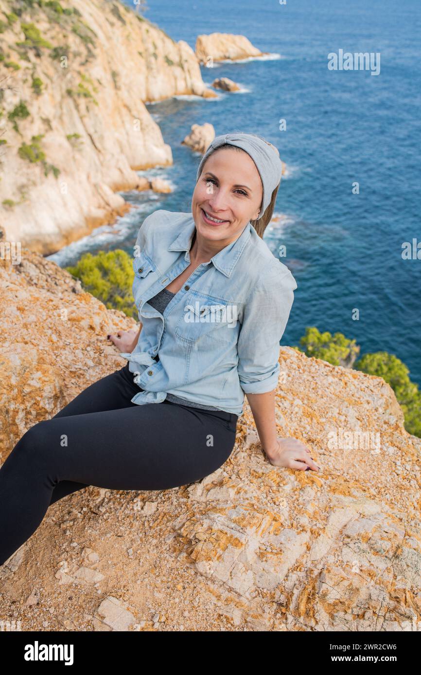 Woman in long sleeves sitting on edge of cliff overlooking Balearic Sea near Tossa de Mar, Spain Stock Photo