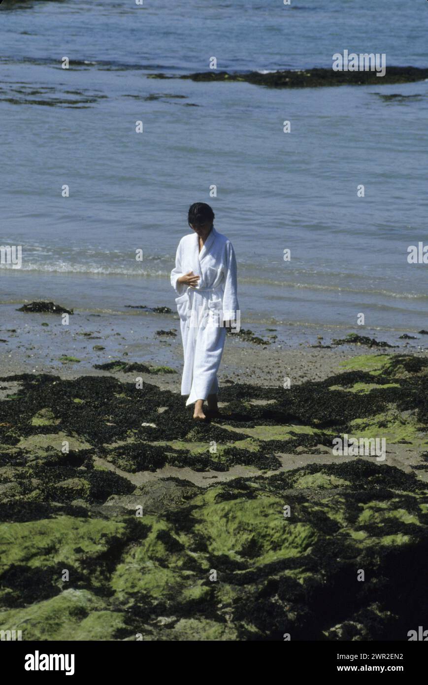 black hair young woman st malo france spa resort thalassotherapy walking on the shore with seagrass in white bath dress Stock Photo