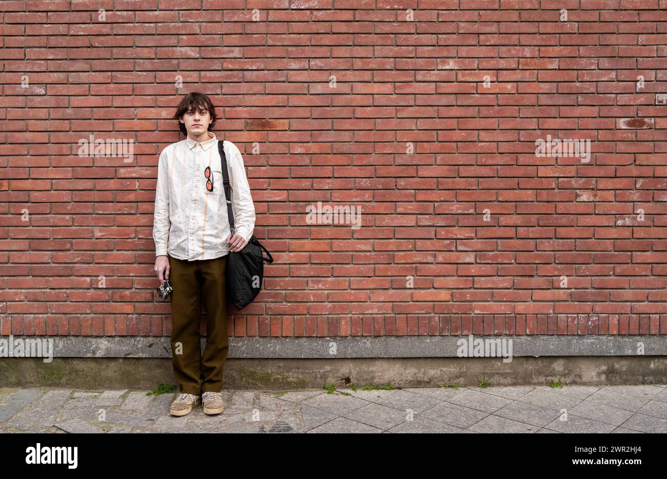 Young man photographer stands with vintage camera against red brick wall, copy space for text. Stock Photo