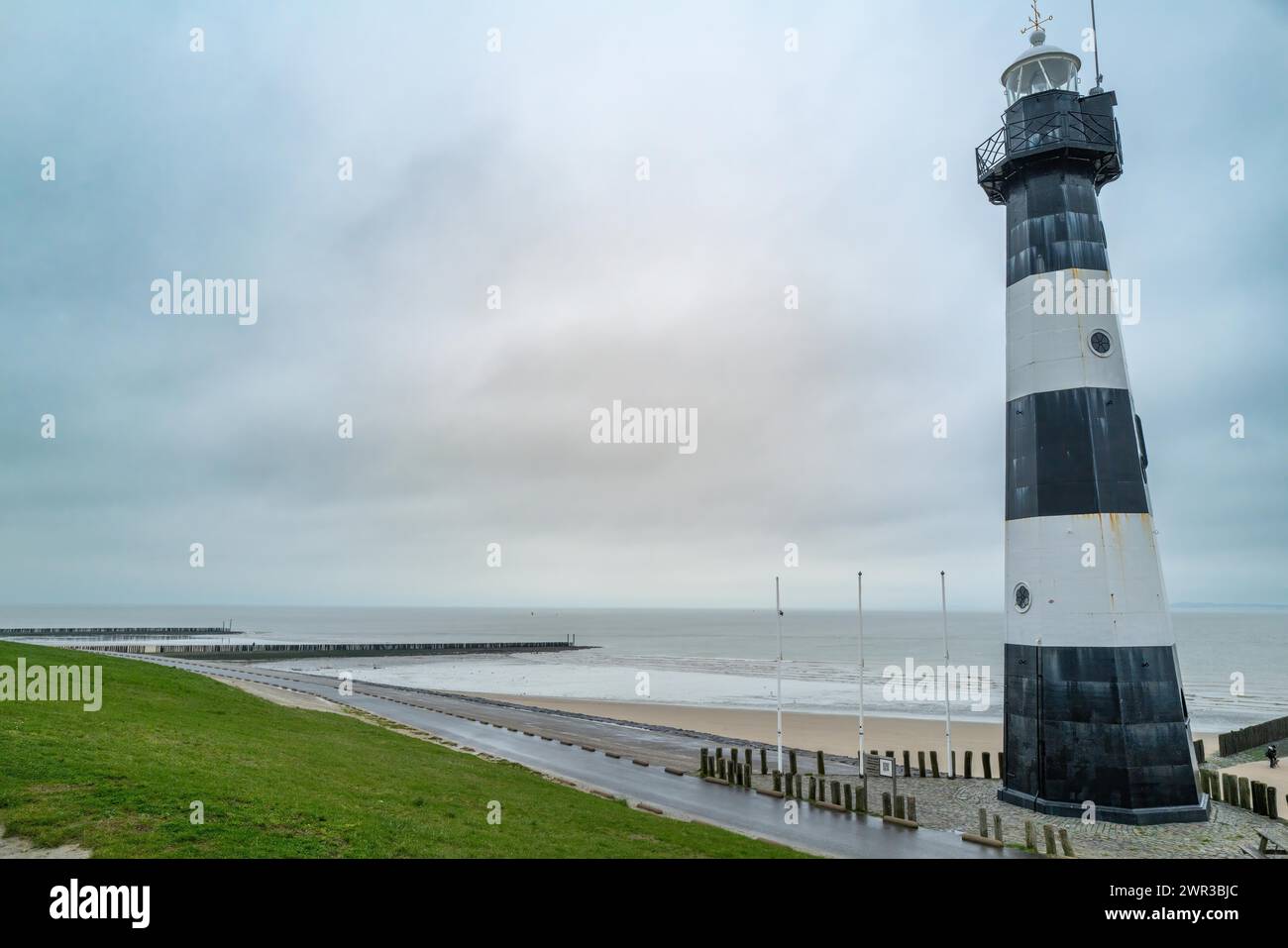 A black and white lighthouse with beach piles and a green area on a cloudy day, Breskens, Zeeland, Netherlands Stock Photo